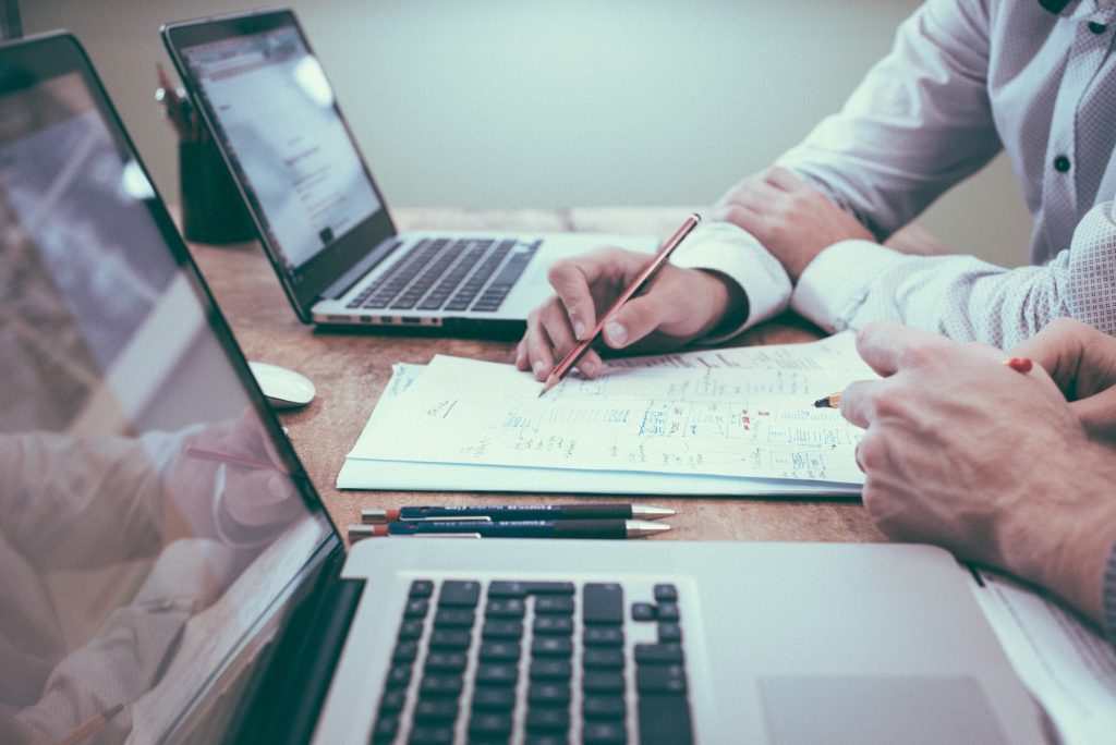 2023 Tech Trends - two/ laptops side by side on a desk with a document in between and the hands of two people sat talking visible. 