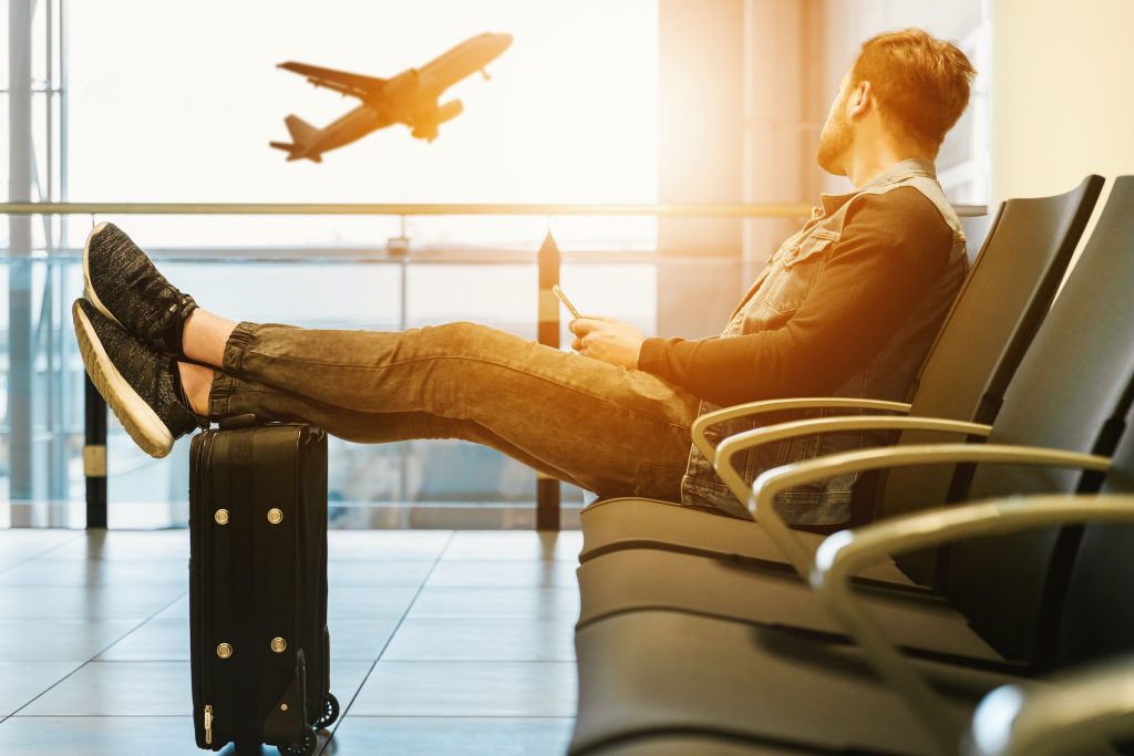 Travel with Tech - man sitting at an airport with a mobile phone in his hand and his feet resting on this case. A aeroplane can be seen taking off in the distance.
